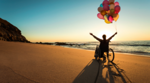 Disability Pride Month: photo of a person in a wheelchair on the beach with their arms in the air holding a bunch of balloons