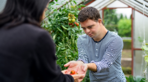 Person with a disability in a greenhouse sharing a tomato with someone else