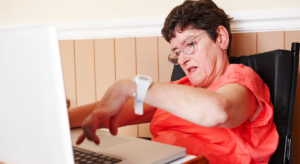 Photo of a white women with a disability in an orange shirt typing at a laptop computer