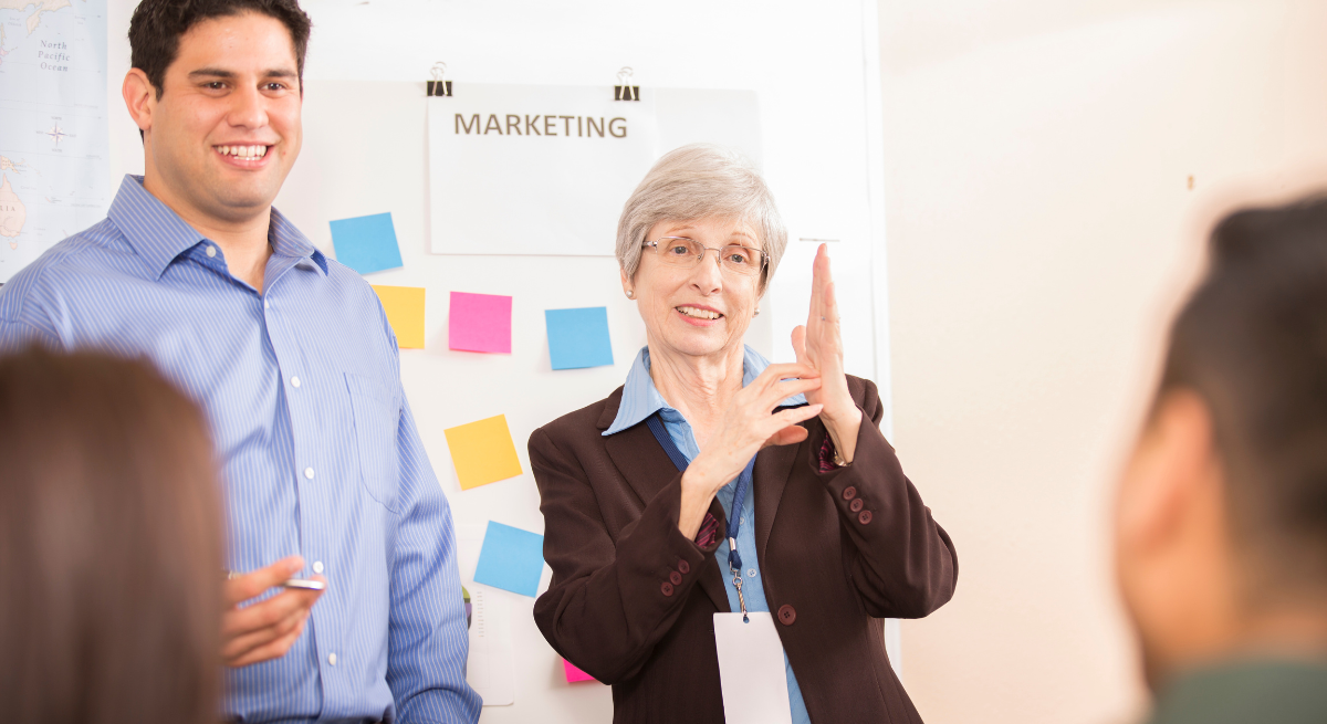 A woman wearing glasses and a suit jacket signs in American Sign Language (ASL) during a meeting, while a man standing beside her smiles. Behind them, colorful sticky notes and a sign reading 'MARKETING' are visible on the wall.