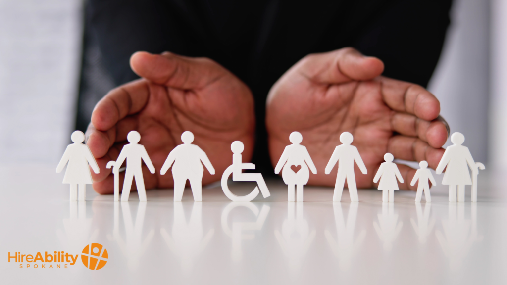 A pair of hands hovers protectively over paper cut-out figures representing diversity, including a person using a wheelchair, elderly figures, pregnant individuals, children, and people of different body types. The logo for HireAbility Spokane is visible in the lower left corner.