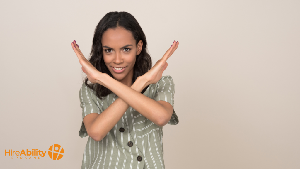 A woman with long dark hair, wearing a green striped shirt, crossing her arms in an 'X' in front of her chest, smiling confidently. The HireAbility Spokane logo is displayed in the lower-left corner.